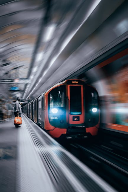 A modern train approaching a station platform with a passenger visible on the left, carrying a small orange suitcase. The train has a sleek, aerodynamic design with a predominantly blue and red exterior, and its headlights are illuminated. The station interior features curved metallic ceilings and blurred motion effects indicating movement. The platform surface appears clean with visible tactile paving along the edge. The background includes part of the station's tunnel-like structure, with some indistinct signage and architectural details. The scene reflects an urban transit environment, relevant to private or alternative transportation methods, and subtly aligns with waste clearance services in terms of efficient, on-site logistical operations supported by structural infrastructure.