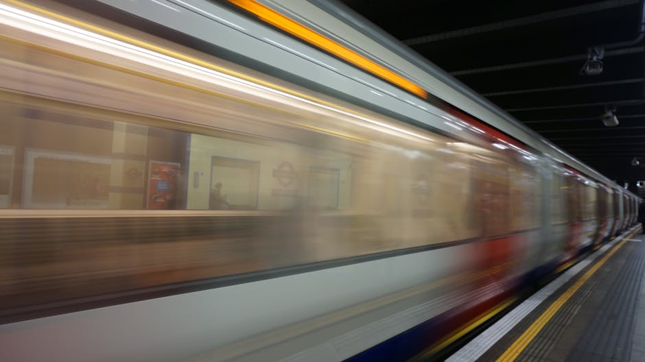 A modern train approaching a station platform with a passenger visible on the left, carrying a small orange suitcase. The train has a sleek, aerodynamic design with a predominantly blue and red exterior, and its headlights are illuminated. The station interior features curved metallic ceilings and blurred motion effects indicating movement. The platform surface appears clean with visible tactile paving along the edge. The background includes part of the station's tunnel-like structure, with some indistinct signage and architectural details. The scene reflects an urban transit environment, relevant to private or alternative transportation methods, and subtly aligns with waste clearance services in terms of efficient, on-site logistical operations supported by structural infrastructure.