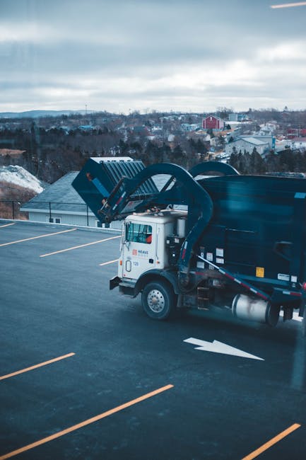 A white and black waste collection truck with a hydraulic arm system is parked on the top level of an outdoor parking structure, with several empty parking spaces visible in the foreground. The truck's side features a small window on the cab, and the hydraulic arm is extended upwards and over the dark blue or black debris container, which appears to be made of metal and has a smooth, glossy finish. The background shows a semi-rural landscape with distant trees, low-rise buildings, and a cloudy sky, suggesting an overcast day. The scene indicates the vehicle may be involved in private waste collection or on-site clearance, possibly supporting independent rubbish removal services like those offered by Waste Clearance Pimlico. The overall environment presents a professional setting for rubbish removal operations, emphasizing the vehicle's role in efficient waste management in an urban or suburban area.