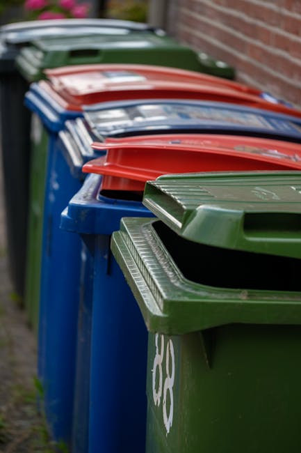 A row of multiple large wheelie bins lined up along a curb beside a brick wall, with the closest bin being green and the others alternating in red, blue, and green colours. The bins are made of durable plastic with textured tops and fitted with lids that are slightly open or closed, revealing some of the interior space. The bins appear clean and weathered, with the green bin showing a white recycling symbol on its side. The background includes an out-of-focus section of pavement and greenery, with natural daylight illuminating the scene evenly. This image reflects a typical setting for private waste collection or on-site rubbish disposal services, highlighting the variety of bins used for different waste streams, often managed independently from local authority collection. Waste Clearance Pimlico provides comprehensive rubbish removal services, including the collection of these wheeled containers, emphasizing their role in efficient waste management.