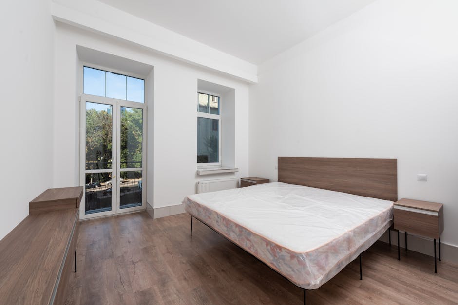 A minimalistic bedroom with white painted walls and a wooden floor, featuring a double bed with a simple wooden headboard and a white mattress, positioned centrally against the right wall. To the left, a large glass door with a white frame leads to an outdoor balcony or terrace, allowing natural light to illuminate the room and revealing a partial view of trees outside. Next to the door, a smaller window with a similar white frame is set into the wall, also bringing in daylight. Opposite the bed, on the far wall, a narrow wooden sideboard is situated, matching the bedside tables on either side of the bed, both with black metal legs and light wood tops. The room has a clean, uncluttered appearance, with no visible furnishings or decorations, emphasizing the space’s simplicity and natural light, which aligns with the context of private or independent waste management or relocation scenarios where minimal furniture is involved, as showcased in the image related to rubbish removal services by Waste Clearance Pimlico.