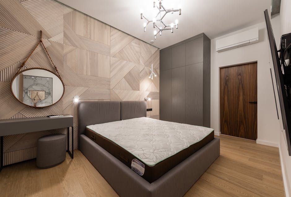A modern bedroom featuring a large bed with a cushioned grey fabric headboard and a white mattress. To the left of the bed, there is a small grey vanity table with a matching round stool beneath it. A round mirror with a thin wooden frame is mounted on the wall above the vanity, reflecting part of a marble surface. The wall behind the bed is decorated with a geometric pattern of light wood panels, adding texture and visual interest to the space. On the right side, there is a closed wooden door with a dark handle, and a sleek grey wardrobe with a smooth finish occupies the corner next to the door. A white wall-mounted air conditioning unit is positioned above the door. The ceiling features a contemporary metallic chandelier with multiple exposed bulb fixtures, providing ample lighting. The flooring is light wood, contributing to the room’s warm and neutral aesthetic. The overall scene depicts a tidy, well-furnished interior suitable for residential living, with no visible clutter or rubbish, illustrating a clean environment where waste clearance services such as those provided by Waste Clearance Pimlico could be relevant for maintaining tidy living spaces.