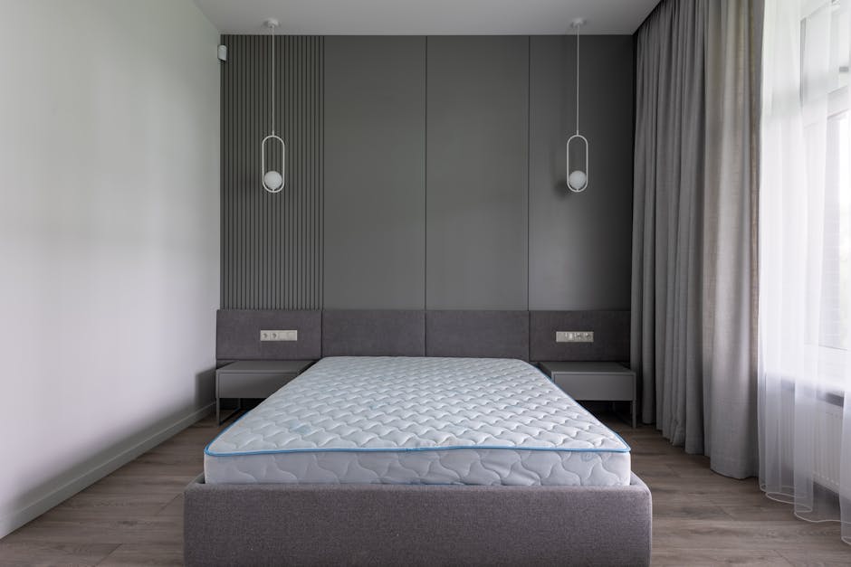 A minimalistic bedroom with white painted walls and a wooden floor, featuring a double bed with a simple wooden headboard and a white mattress, positioned centrally against the right wall. To the left, a large glass door with a white frame leads to an outdoor balcony or terrace, allowing natural light to illuminate the room and revealing a partial view of trees outside. Next to the door, a smaller window with a similar white frame is set into the wall, also bringing in daylight. Opposite the bed, on the far wall, a narrow wooden sideboard is situated, matching the bedside tables on either side of the bed, both with black metal legs and light wood tops. The room has a clean, uncluttered appearance, with no visible furnishings or decorations, emphasizing the space’s simplicity and natural light, which aligns with the context of private or independent waste management or relocation scenarios where minimal furniture is involved, as showcased in the image related to rubbish removal services by Waste Clearance Pimlico.