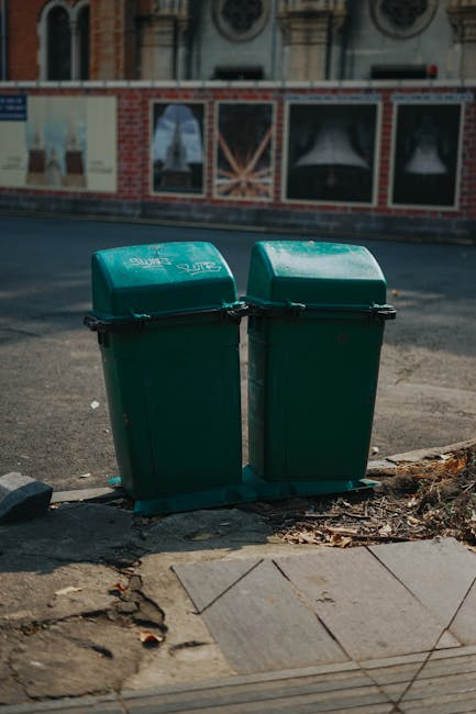 Two green wheeled rubbish bins with closed lids positioned side by side on a pavement at the edge of a street. The bins are made of hard plastic with a textured surface, and each lid has a slight curve with a lid hinge mechanism visible along the front edge. They sit on a black plastic base that raises them slightly above the ground. Surrounding the bins, the pavement is made of concrete tiles, some of which appear cracked and uneven, with scattered dry leaves and debris nearby. In the background, there is a street with asphalt surface, and further back, a decorative brick wall with large window frames and painted murals or advertisements. The scene is illuminated by natural daylight, creating minimal shadows, and the environment suggests an urban setting where private waste collection or rubbish removal services, such as Waste Clearance Pimlico, might be involved in on-site clearance or alternative waste handling outside local authority disposal channels.