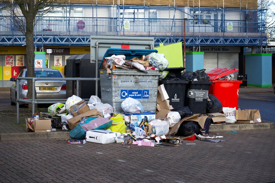 A concentrated pile of mixed waste, including cardboard boxes, paper bags, plastic bags, and various crumpled packaging materials, is gathered on a paved sidewalk in an urban environment. The debris consists of both flattened and unflattened cardboard packaging, several black and red garbage bags, and loose paper and plastic items. Behind the trash, there are black and gray recycling bins, some partially open, with lids either lifted or tilted to reveal contents, and a large gray recycling container labeled for mixed paper and cardboard. The scene is set against a background of a store façade with bright signage, a parked silver car on the left, and a building under construction covered with blue scaffolding and netting, suggesting nearby renovation activity. Natural daylight casts shadows across the area, indicating an outdoor setting, with a metal railing bordering the pavement. The overall scene exemplifies a typical urban rubbish accumulation and disposal context, often associated with waste clearance services focused on private or alternative waste collection, as provided by Waste Clearance Pimlico.