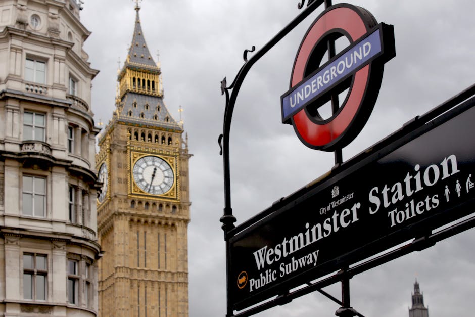 A close-up shot of a black and white signboard for Westminster Station, a part of the London Underground, positioned in the foreground. The sign displays directions for public subway access and toilets, with icons indicating pedestrian pathways. Above the sign, an iconic roundel with a red circle and blue bar labeled 'UNDERGROUND' is visible. In the background, the Elizabeth Tower, commonly known as Big Ben, is prominent with its ornate, gold-accented clock face showing the time as approximately 2:55. The tower's Gothic architectural details and spire extend upward, with the building's beige stone façade showing intricate craftsmanship and texture. The sky above is overcast with gray clouds, creating a subdued lighting atmosphere typical of a cloudy day in central London. This urban scene combines elements of public transportation infrastructure with historic architectural landmarks, illustrating typical surroundings encountered during city waste management and maintenance activities at busy transport hubs, such as the work carried out by waste clearance services. The environment suggests a well-known iconic area where independent waste handling may be necessary alongside communal facilities.