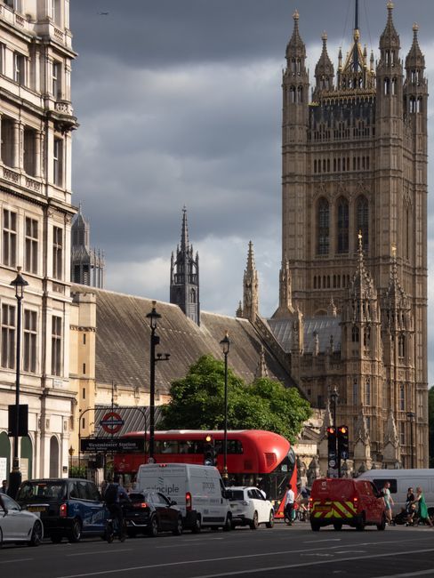 A busy street scene in Westminster with a view of the iconic Palace of Westminster and its prominent clock tower, known as Big Ben, in the background under a partly cloudy sky. In the foreground, there are several vehicles, including a white delivery van, a red double-decker bus, a small red utility vehicle with reflective stripes, and a few cars parked along the curb. Pedestrians are visible walking across the street and waiting at the crosswalk. The surrounding area includes historic buildings with detailed stone facades and decorative architectural elements, as well as street lamps and traffic signals. The image captures an urban environment typical of central London, illustrating the mix of traffic, pedestrian activity, and historic landmarks that may relate to alternative waste disposal via private rubbish collection or on-site clearance, as offered by waste management services like Waste Clearance Pimlico.