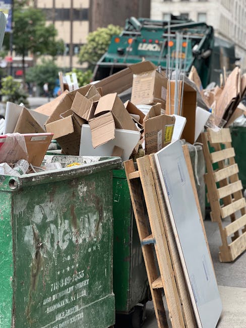 A large green waste disposal skip filled with a variety of cardboard boxes, both flattened and partially assembled, alongside some white packaging containers, situated on an urban street. The boxes are of different sizes and materials, with some showing printed labels and tape. Several wooden pallets, used for supporting and transporting the waste, lean against the side of the skip. In the background, there is a street scene with blurred trees, buildings, and a green waste collection vehicle, indicating this scene relates to private waste handling and rubbish removal services. The area appears to be a typical city environment with paved surfaces and nearby traffic or parked vehicles. Natural daylight illuminates the scene, subtly highlighting the textures of cardboard and wood, consistent with a waste clearance operation, such as those offered by Waste Clearance Pimlico, for on-site or independent rubbish collection.