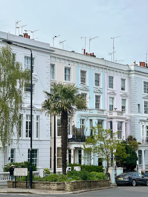 A multi-storey residential building with a white facade featuring classical architectural details, including decorative window mouldings and a small balcony. In the foreground, there is a narrow pavement lined with green shrubbery and small trees, including a tall palm tree situated close to the building. A black street lamp stands near the palm, and a dark grey car is parked on the street beside a low brick wall enclosing a garden area. The building's rooftop is equipped with several TV antennas, and the sky above appears overcast, casting diffuse natural light across the scene. The overall environment suggests a well-maintained urban residential area, typical of Pimlico, with attention to garden landscaping and street furniture, reflecting the local character and private waste handling practices associated with such properties. Waste Clearance Pimlico’s services could be relevant here for managing rubbish removal from residential properties with similar architectural features.
