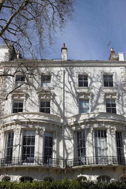 A multi-storey residential building with a white facade featuring classical architectural details, including decorative window mouldings and a small balcony. In the foreground, there is a narrow pavement lined with green shrubbery and small trees, including a tall palm tree situated close to the building. A black street lamp stands near the palm, and a dark grey car is parked on the street beside a low brick wall enclosing a garden area. The building's rooftop is equipped with several TV antennas, and the sky above appears overcast, casting diffuse natural light across the scene. The overall environment suggests a well-maintained urban residential area, typical of Pimlico, with attention to garden landscaping and street furniture, reflecting the local character and private waste handling practices associated with such properties. Waste Clearance Pimlico’s services could be relevant here for managing rubbish removal from residential properties with similar architectural features.