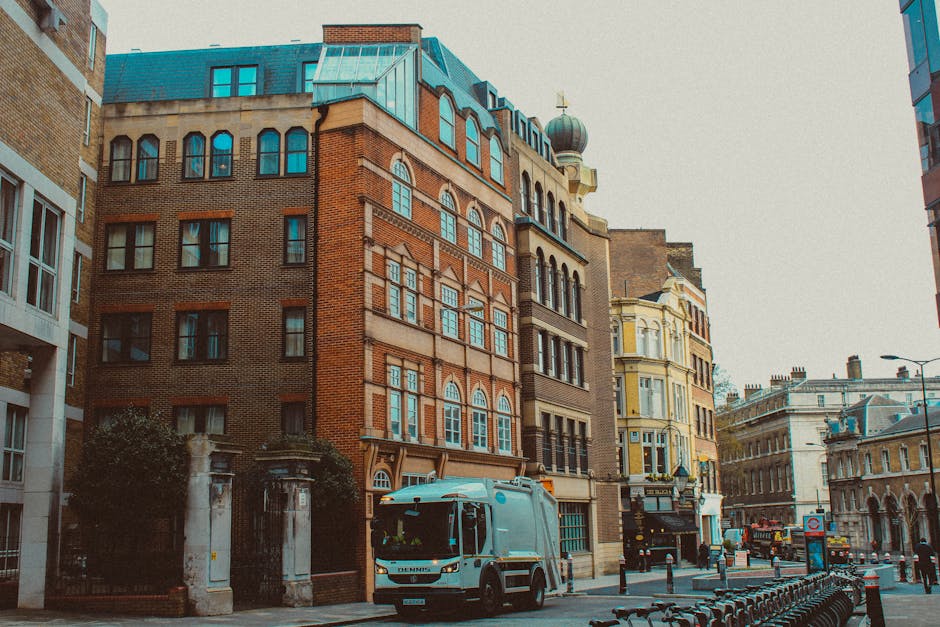 A city street scene showing a compact refuse collection truck parked close to a multi-storey brick building with a blue mansard roof and decorative dormer windows, situated next to a white stone gatepost at the corner of the sidewalk. The truck, featuring a white cab and a metallic silver waste body, is positioned to the left in the foreground, with clear visibility of its front and side windows. The background reveals a range of historic multi-storey buildings, including one with a distinctive green-domed turret and ornate detailing, alongside a pale yellow building with bay windows and decorative cornices, all under an overcast sky that casts diffused light over the scene. The pavement features a line of metal barriers separating the roadside from pedestrians, and a bus and other vehicles are visible further down the street, contributing to the urban environment associated with private waste collection services by Waste Clearance Pimlico, which operates to provide alternative rubbish removal options outside local authority collections.
