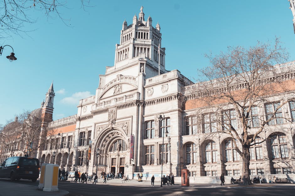 A large, historic stone building with intricate architectural details, including arched windows, decorative carvings, and a prominent central tower topped with a spire, situated on a street corner under a clear blue sky. The façade features a mix of smooth and textured stone surfaces with a light beige tone, and the windows are framed with dark trim. On the sidewalk in front, both pedestrians and vehicles are visible, and leafless trees with spreading branches line the street, indicating a winter or early spring season. Street lamps and a British telephone box are also present nearby. The environment appears urban and well-maintained, with the building likely serving as a cultural or civic landmark. The scene subtly reflects the context of private or alternative waste management, suitable for discussions about independent waste clearance or specialist rubbish removal services, aligned with the theme of rubbish removal in a professional, neutral tone that supports SEO relevance for waste clearance in the area.