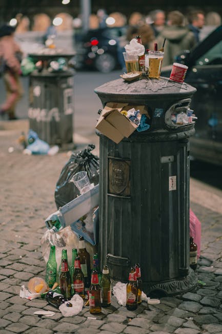 A black, cylindrical metal waste bin with a domed lid is situated on a paved street surface in an urban environment, surrounded by scattered litter including glass bottles, paper, and plastic waste primarily concentrated at its base and on top. The bin's surface shows some signs of weathering and minor rust, with various small items such as a cardboard box, used paper cups, and drink containers placed on the lid, some of which have spilled over onto the ground. In the background, blurred figures of pedestrians and additional parked vehicles are visible, indicating a busy public area. The scene appears to depict informal rubbish accumulation, possibly from informal disposal rather than organized waste collection, aligning with the context of private waste handling or unmanaged rubbish and illustrating the importance of proper waste management services, such as those offered by Waste Clearance Pimlico, in keeping urban spaces tidy. The overall atmosphere is one of casual neglect, with environmental and cleanliness considerations relevant to rubbish removal services.