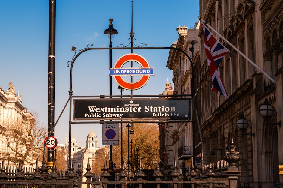 A street scene outside Westminster Station in London featuring a large, circular red and blue underground sign suspended from a decorative black metal arm attached to a street lamp. Beneath the sign is a rectangular black and white information board indicating directions to the public subway and toilets, with the words 'Westminster Station' prominently displayed. To the right, a Union Jack flag is mounted on a pole attached to an ornate, historic building with stone facades and arched windows. The background shows clear blue sky, along with additional historic buildings and trees with bare branches, suggesting late autumn or winter. The image captures the urban environment surrounding Westminster underground station, highlighting the classic London street aesthetic, with a focus on transportation signage and nearby architecture, relevant to visitors seeking public transport and local amenities in the Westminster area. Waste Clearance Pimlico provides services that include handling various types of rubbish removal in such environments, supporting alternative waste management options for residents and businesses in the district.