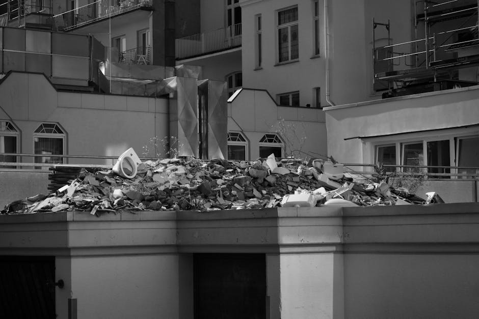 A large pile of construction debris and rubbish is situated on a rooftop ledge in an urban setting. The debris includes broken concrete, plaster, and assorted fragments of building materials, predominantly in shades of grey and white, with some darker pieces. The pile appears unstable, with scattered debris falling over the edge of the ledge. In the background, modern multi-storey buildings with white facades, rectangular windows, and balconies are visible, along with scaffolding on the right side, indicating ongoing construction or renovation work. The scene is illuminated by natural daylight, rendering sharp shadows and fine details of the rubble and surrounding structures. The image captures an example of private or on-site waste accumulation, which companies like Waste Clearance Pimlico might handle as part of their rubbish removal services for renovation waste management in urban environments.