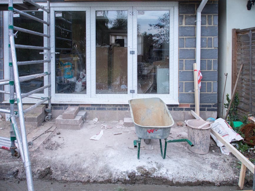 The image depicts a residential outdoor area in the midst of construction or renovation, with a concrete patio space directly in front of a set of large, double-glazed sliding glass doors. To the left, there's metal scaffolding with horizontal bars, indicating ongoing work on the building exterior. The ground surface appears uneven and dusty, likely impacted by construction debris, with scattered, crumpled paper or packaging. In the foreground, a yellowed, worn wheelbarrow with a metal frame and plastic or fiberglass basin is positioned centrally, surrounded by various construction materials and tools, including a large plastic bucket, a piece of wood, and some small hand tools. The scene is framed by brick and wooden fencing on the right, along with a tall, slender wooden support or pipe tied with red-and-white caution tape, suggesting ongoing work or temporary safety measures. Inside the building, the reflection in the glass shows some furniture or interior items, though details are unclear. The overall scene looks like a private property undergoing renovation, with Waste Clearance Pimlico occasionally involved in managing the disposal of building waste or excess materials, typical of private rubbish removal or alternative waste handling options.